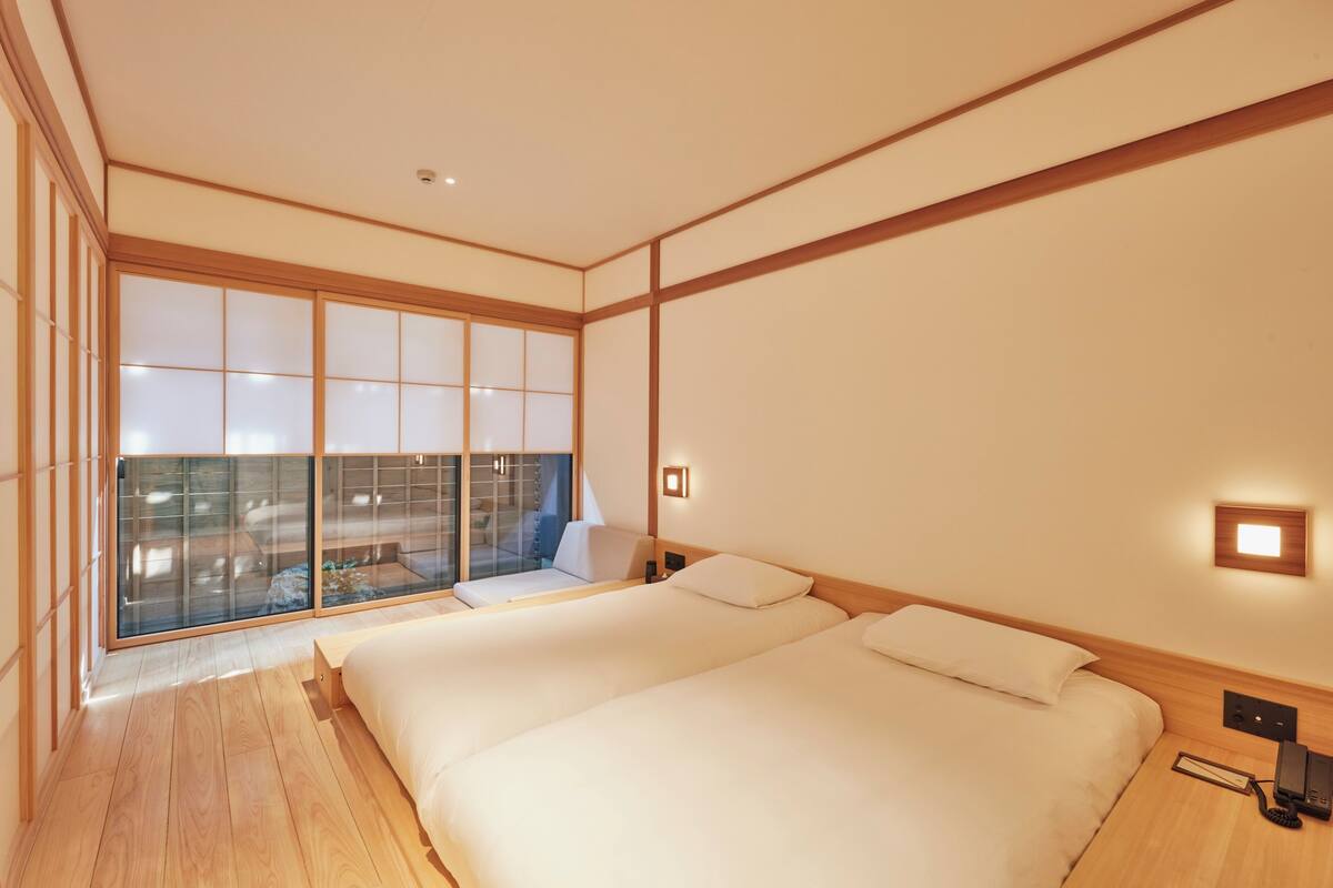 A hotel room with two beds, a large window with a view, wooden flooring, and a small bedside table with a phone.