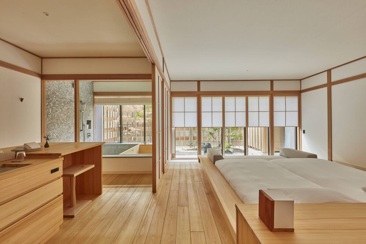 A modern Japanese-style bedroom with a wooden floor, a large bed, a wooden desk, and a view of a garden through large windows.