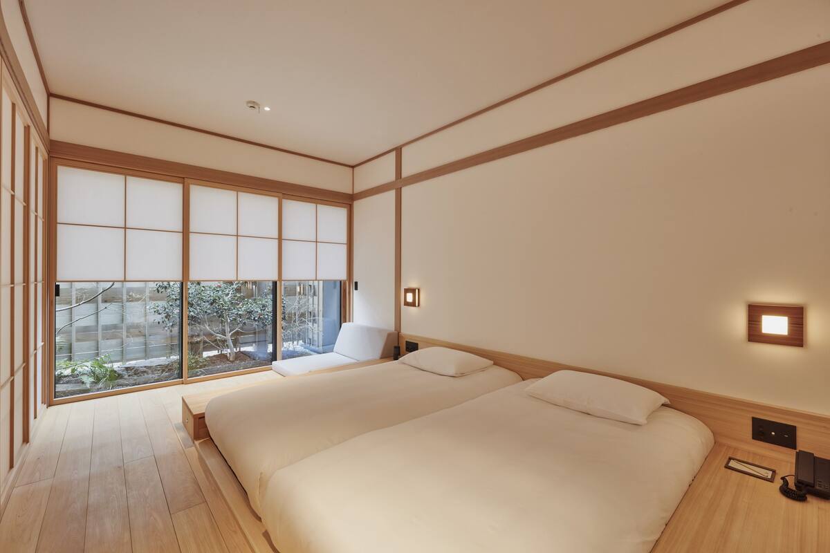 A modern hotel room with two beds, a large window, and a view of the outdoors.