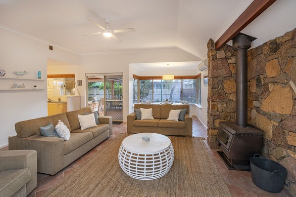 A living room with a stone fireplace, a ceiling fan, and a view of the outdoors through large windows.