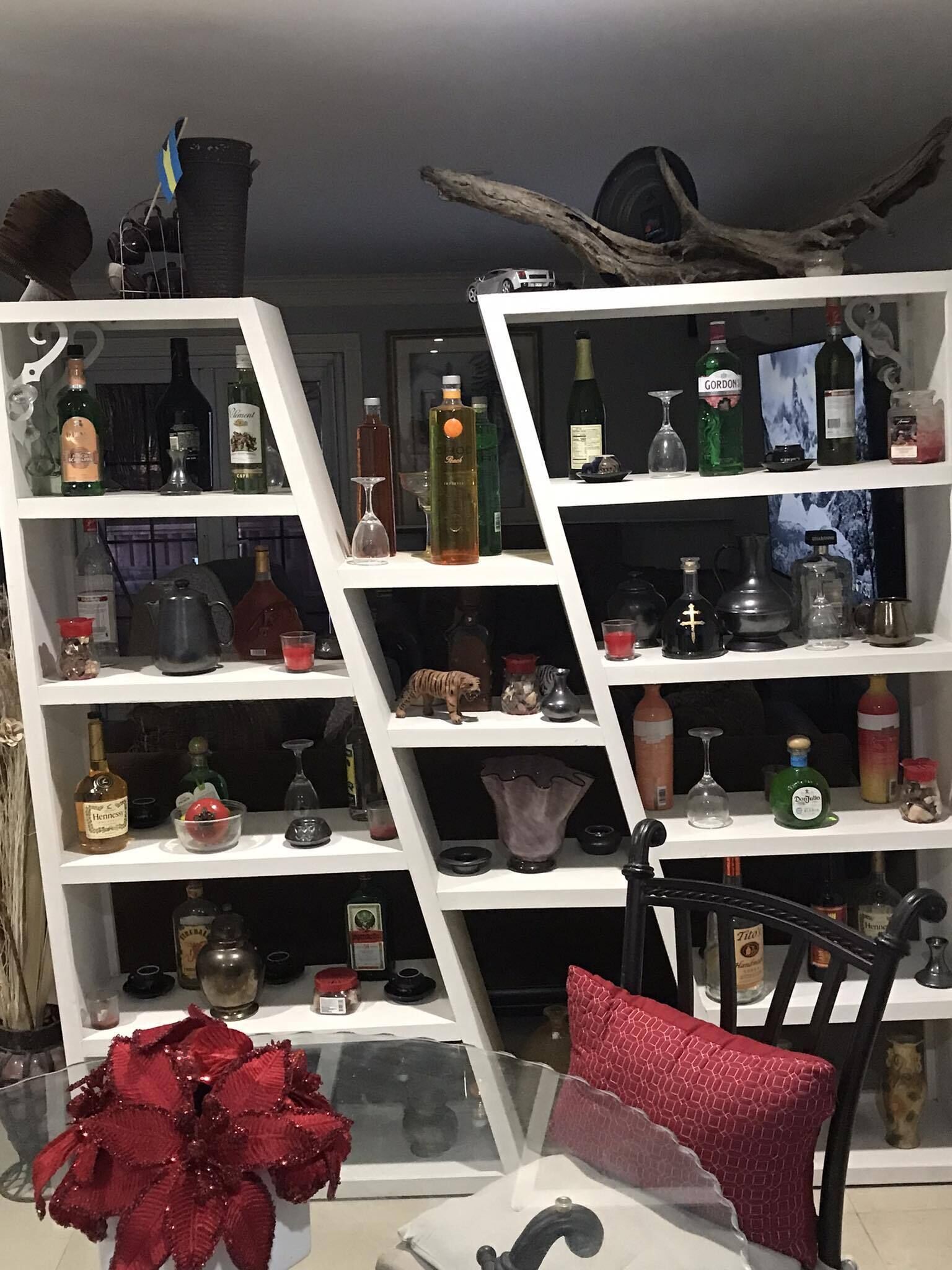 A room with a white shelving unit filled with various bottles and glassware, a red decorative plant, and a black chair with a red cushion.