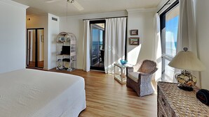 Une chambre d’hôtel avec un lit, un ventilateur de plafond, une chaise en osier, une petite table avec une lampe et une vue sur l’océan par une grande fenêtre.