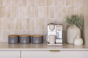 Een keukenblad met drie grijze bakken, gelabeld met thee, suiker en koffie, een koffiezetapparaat en een witte vaas met gedroogde planten.
