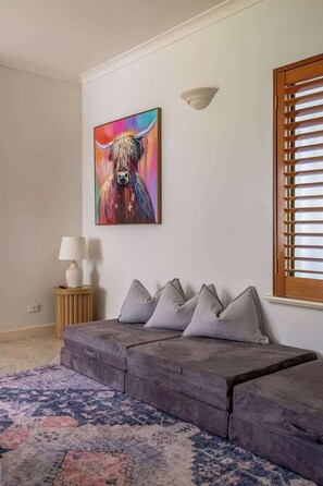 A modern living room with a grey sofa, a colorful painting, a lamp, and a window with blinds.