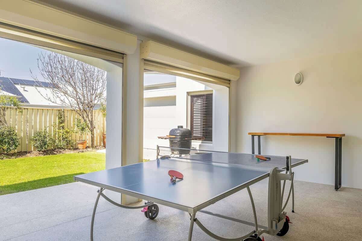 A ping pong table in a room with large windows, a barbecue grill outside, and a wooden table to the right.