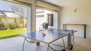 A ping pong table in a room with large windows, a barbecue grill outside, and a wooden table to the right. - Torretta by the Bay (Dunsborough)