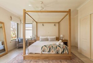 A four-poster bed with a wooden frame, a bedside table with a lamp, and a view of the outdoors through a window.