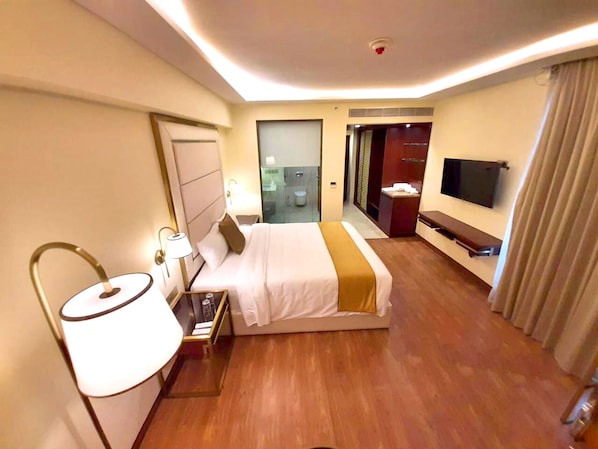 A hotel room with a bed, bedside table, lamp, TV, and a bathroom visible through a glass door.