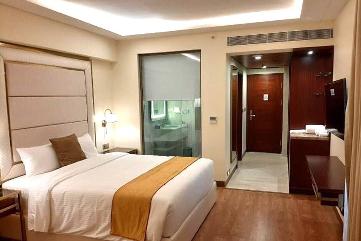 A hotel room with a large bed, a bedside table, a desk, a mirror, and a door leading to a bathroom.