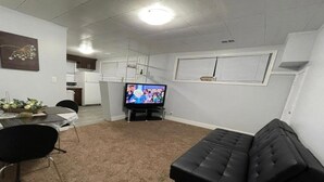 Un espacio habitable compacto con comedor, un sofá negro, televisión y cocina al fondo.