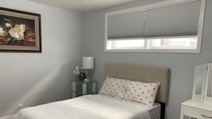 Un dormitorio con cama, mesita de noche, lámpara, ventana con persianas y un cuadro en la pared.