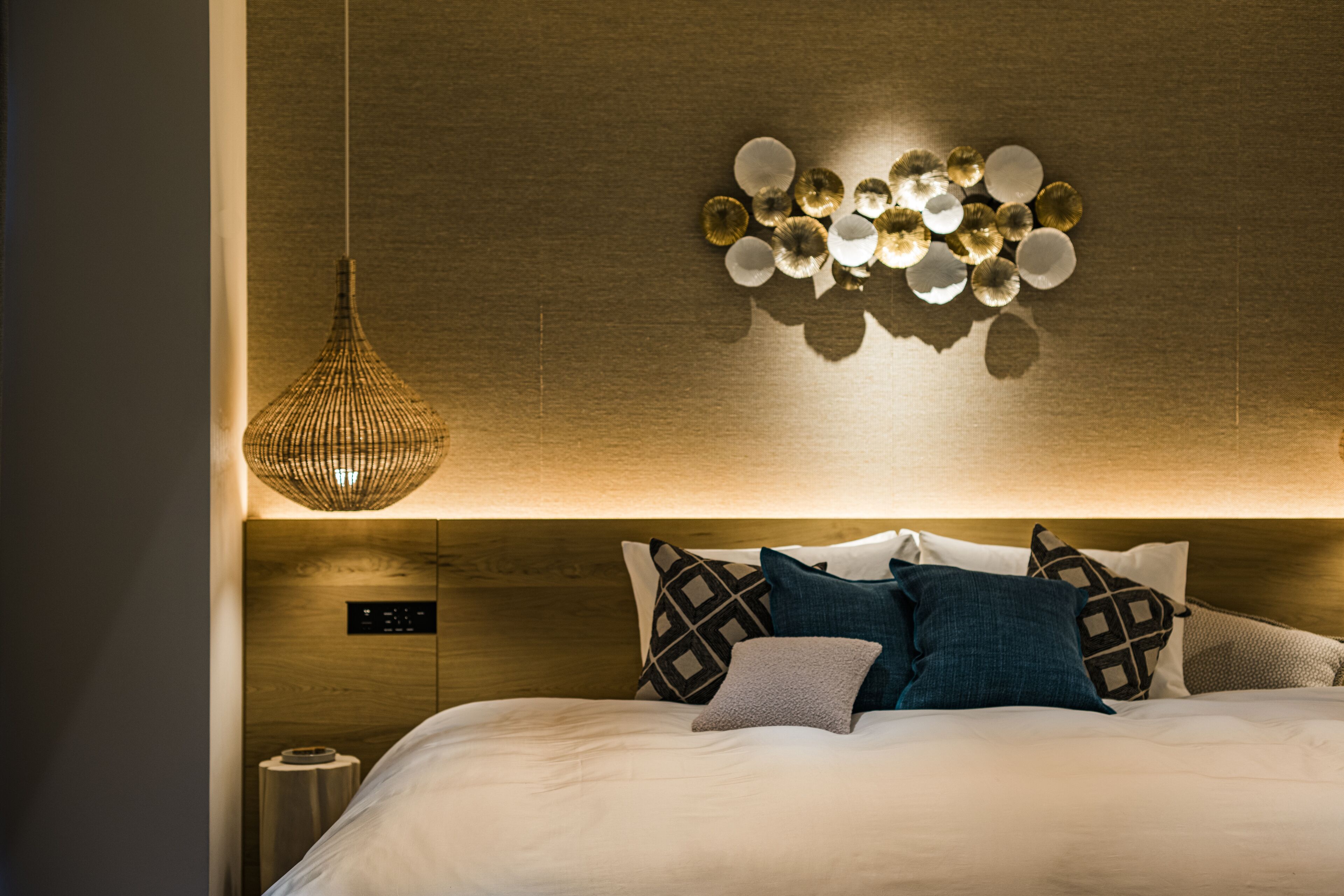 A neatly made bed with patterned pillows, a hanging light fixture, and a wall-mounted control panel.