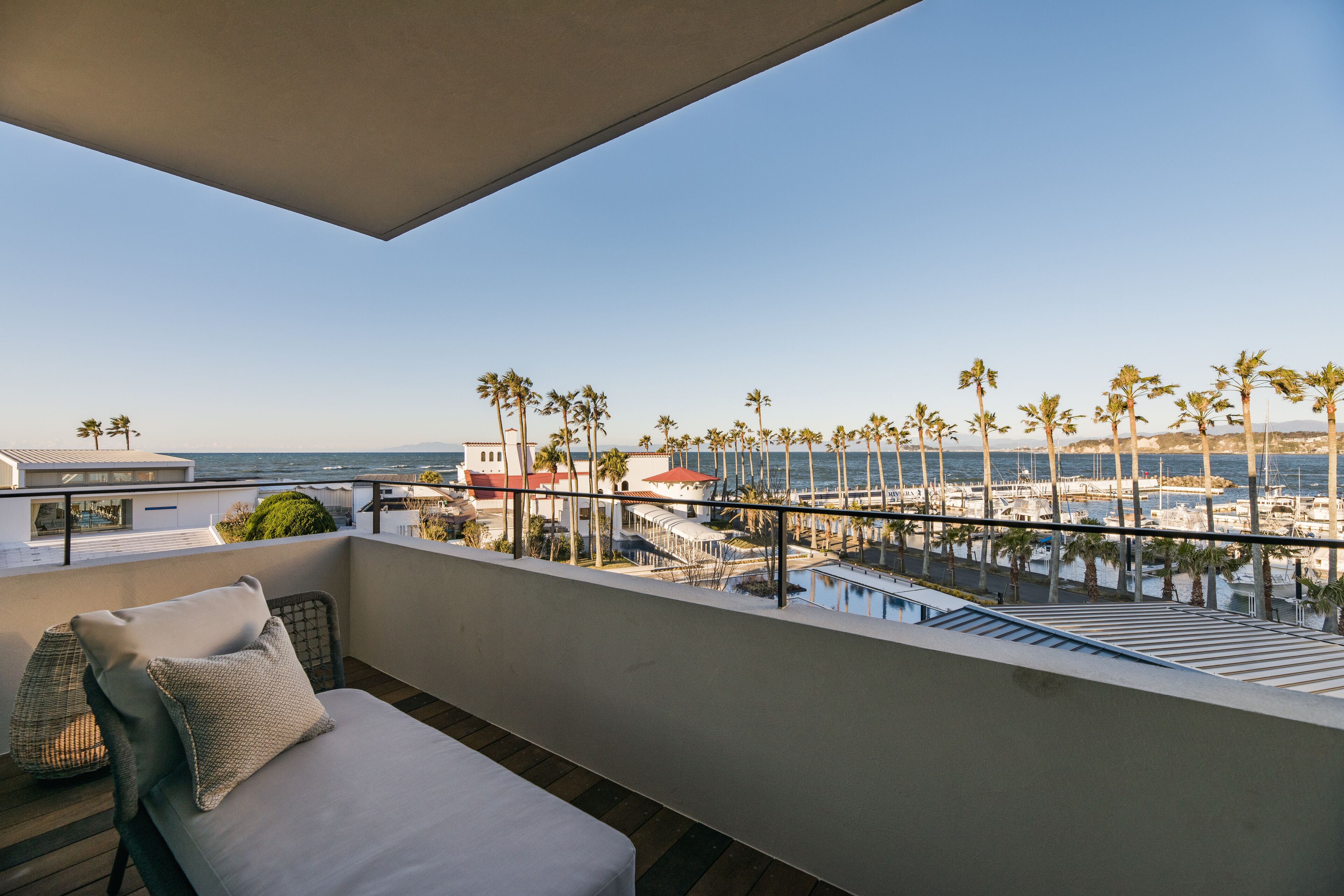A balcony with a view of the sea, palm trees, and a building with a red roof.