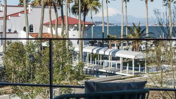 A balcony with a view of palm trees, a building with a red roof, and a body of water in the distance.