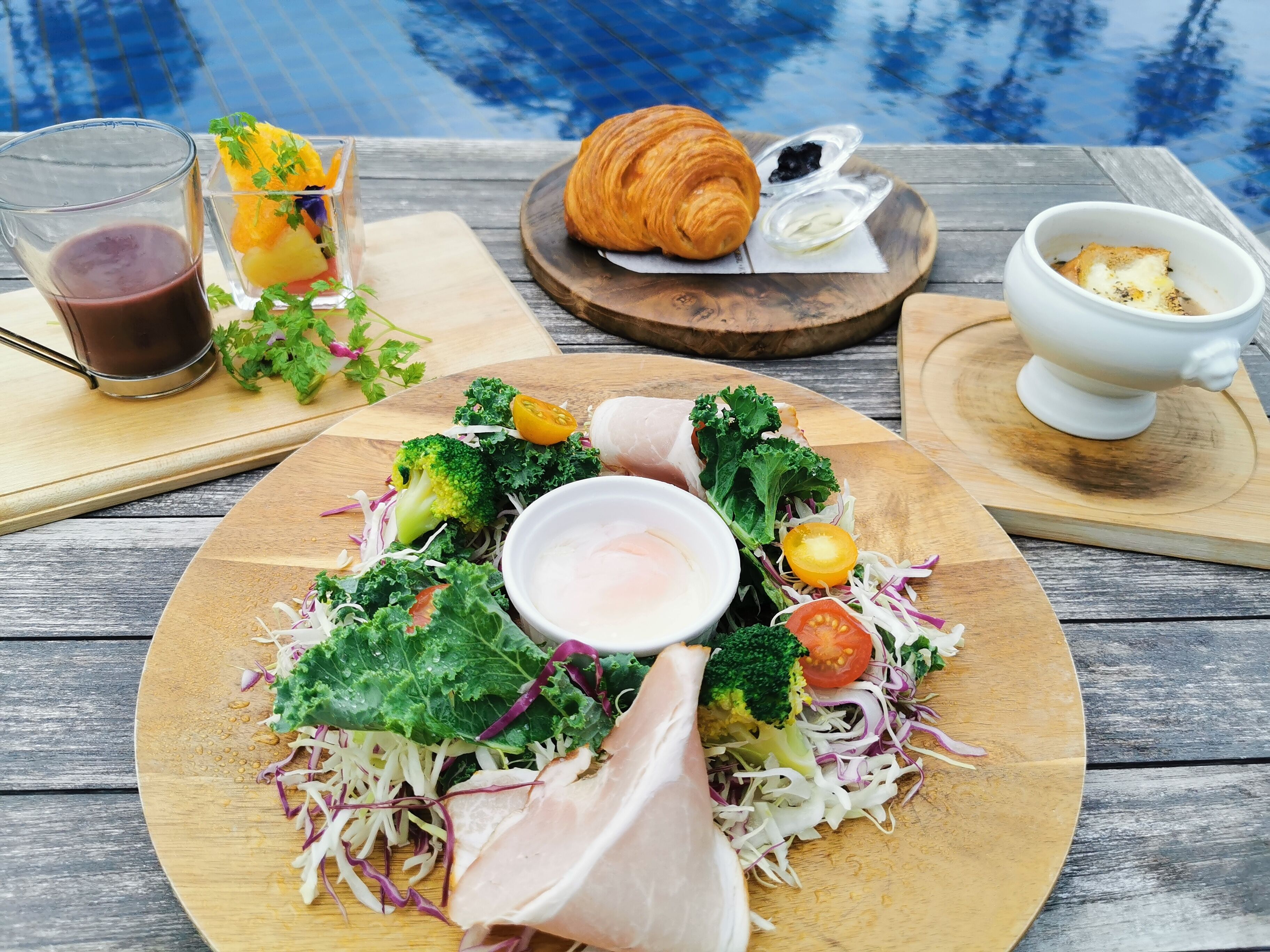 A wooden table with a meal consisting of a salad, a croissant, a glass of juice, and a bowl of soup.