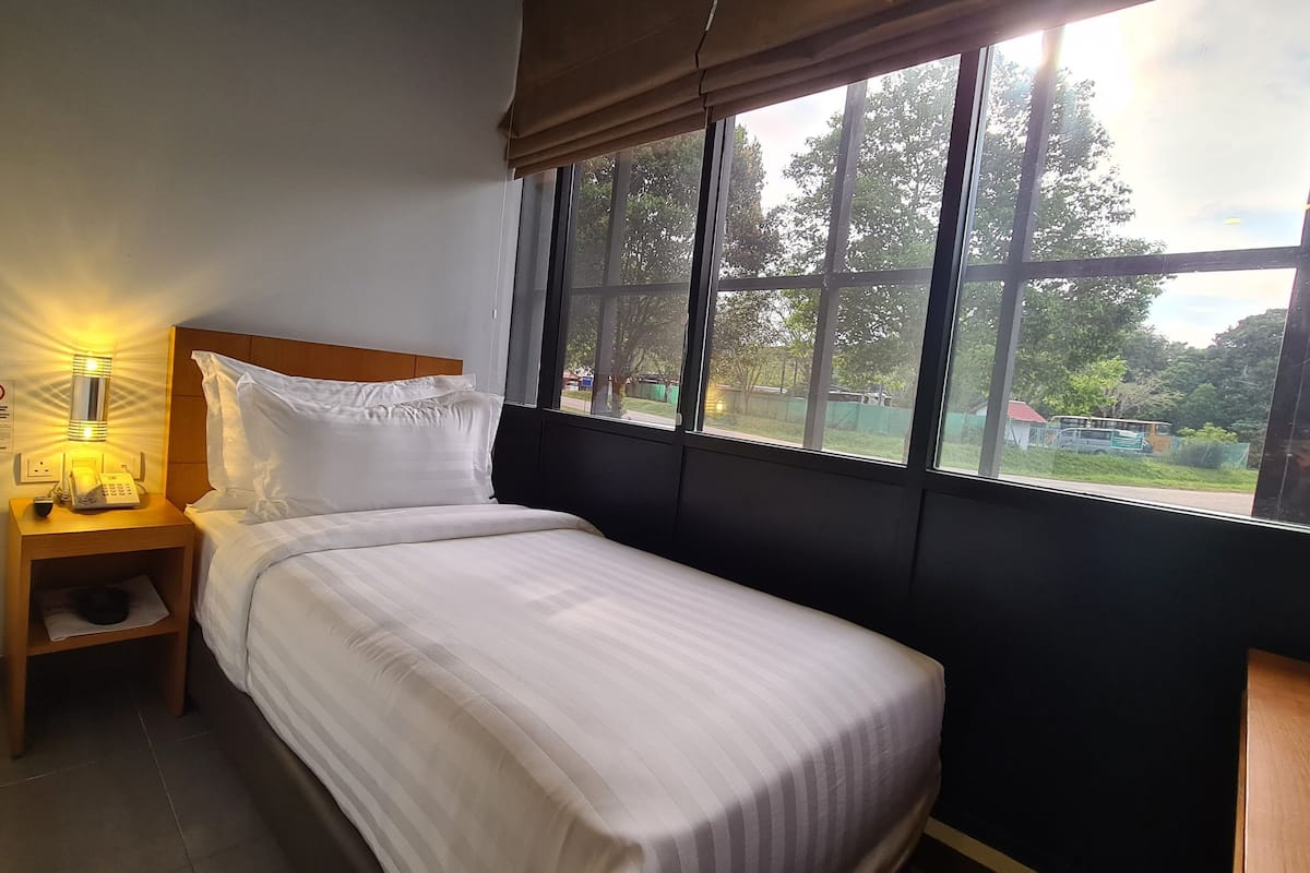 Quarto pequeno com uma cama de solteiro, cabeceira de madeira, mesa de cabeceira com abajur e janela com vista para o exterior.