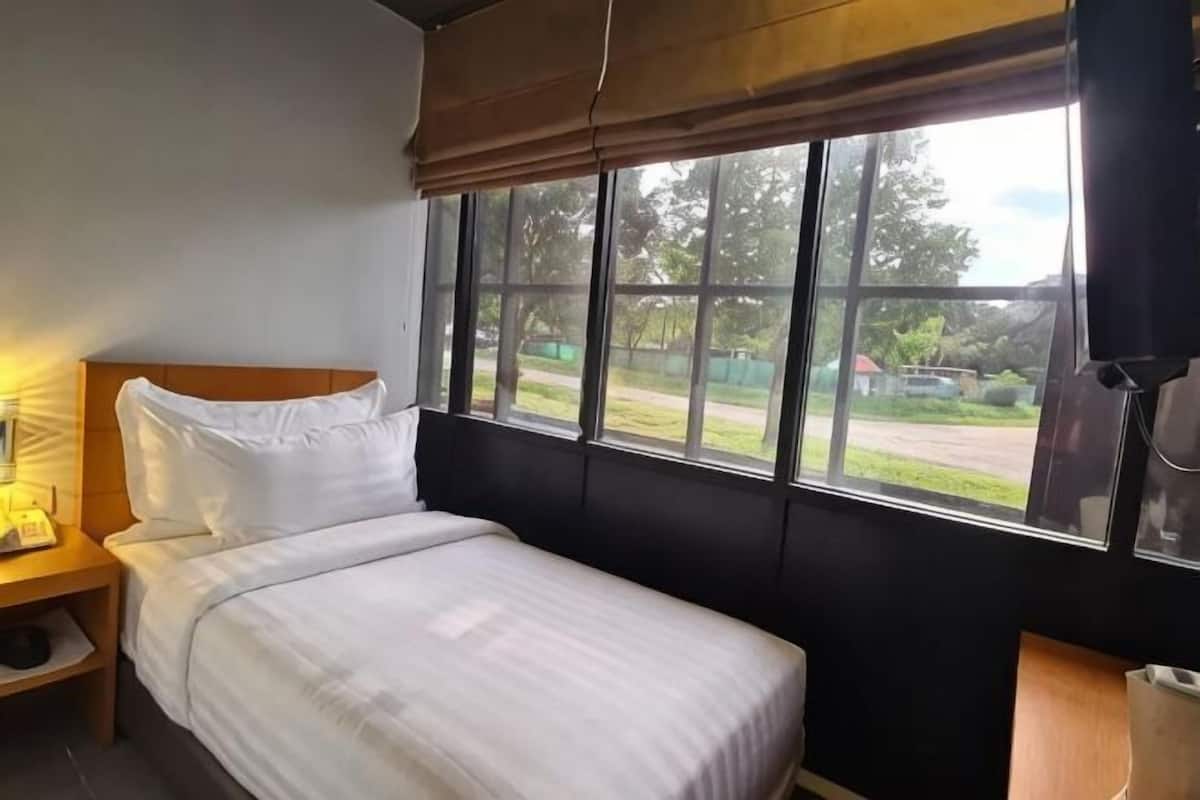 Uma cama de solteiro com cabeceira de madeira, uma mesinha de cabeceira com abajur, e uma janela com vista para o exterior.