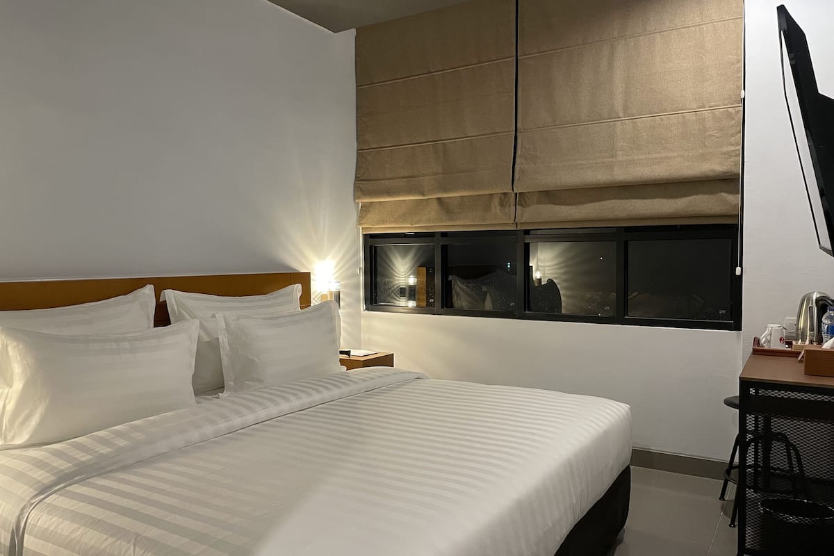 Quarto de hotel com uma cama grande, uma escrivaninha com cadeira, uma janela com persianas e uma televisão de tela plana fixada na parede.