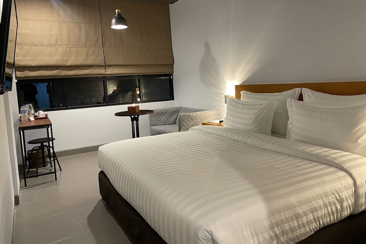 Quarto de hotel com uma cama grande, uma mesa pequena com uma cadeira, uma janela com vista e uma luminária no teto.