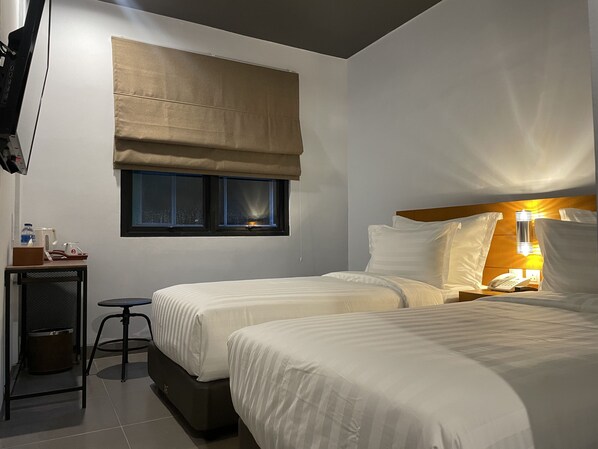 A hotel room with two beds, a small table with a chair, a TV mounted on the wall, and a window with a view of the city at night.