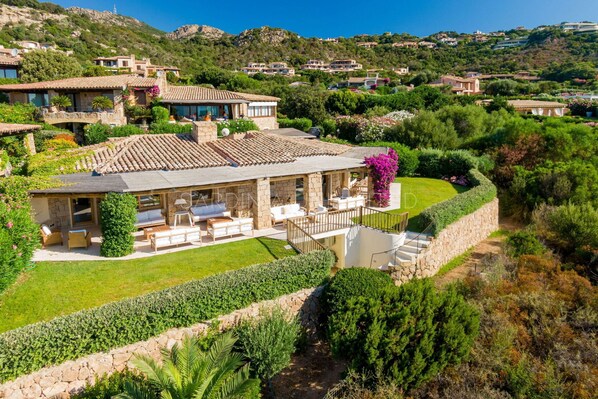 A Mediterranean-style villa with a terracotta-tiled roof, a covered patio with outdoor furniture, and a lush garden.