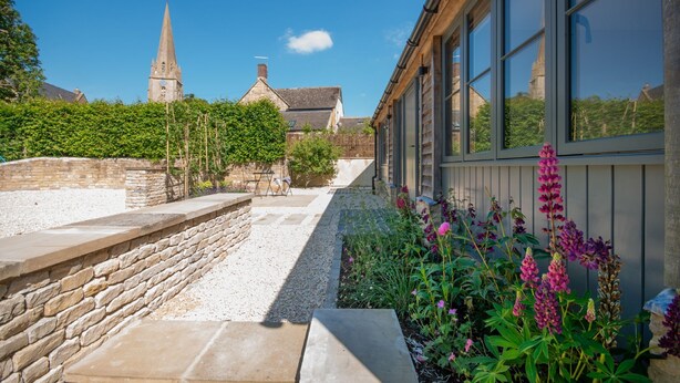 A stone pathway leading to a building with large windows, a stone wall, and a garden with flowers. - Three bedroom Cotswold holiday cottage - Steeple View (Todenham)