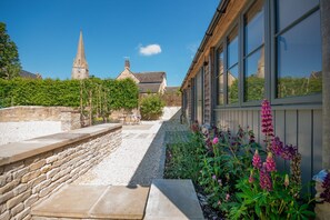 Un camí de pedra que condueix a un edifici amb finestres grans, una paret de pedra i un jardí amb flors.