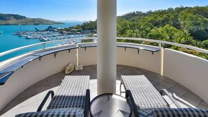 A balcony with two checkered lounge chairs and a small table, overlooking a marina with boats and a lush green landscape.