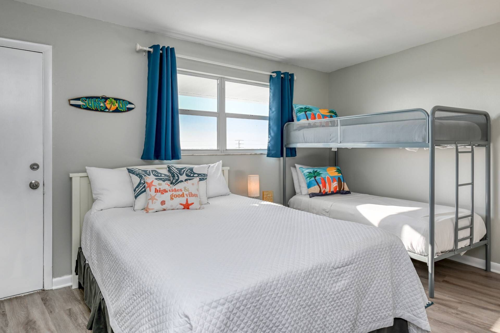 A bedroom with a bunk bed, two single beds, a window with curtains, and a surfboard decoration.
