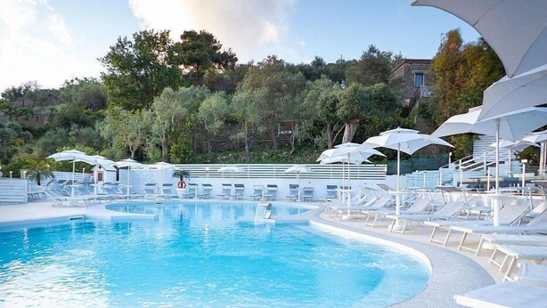 A swimming pool with white lounge chairs and umbrellas, surrounded by trees and a building in the background.