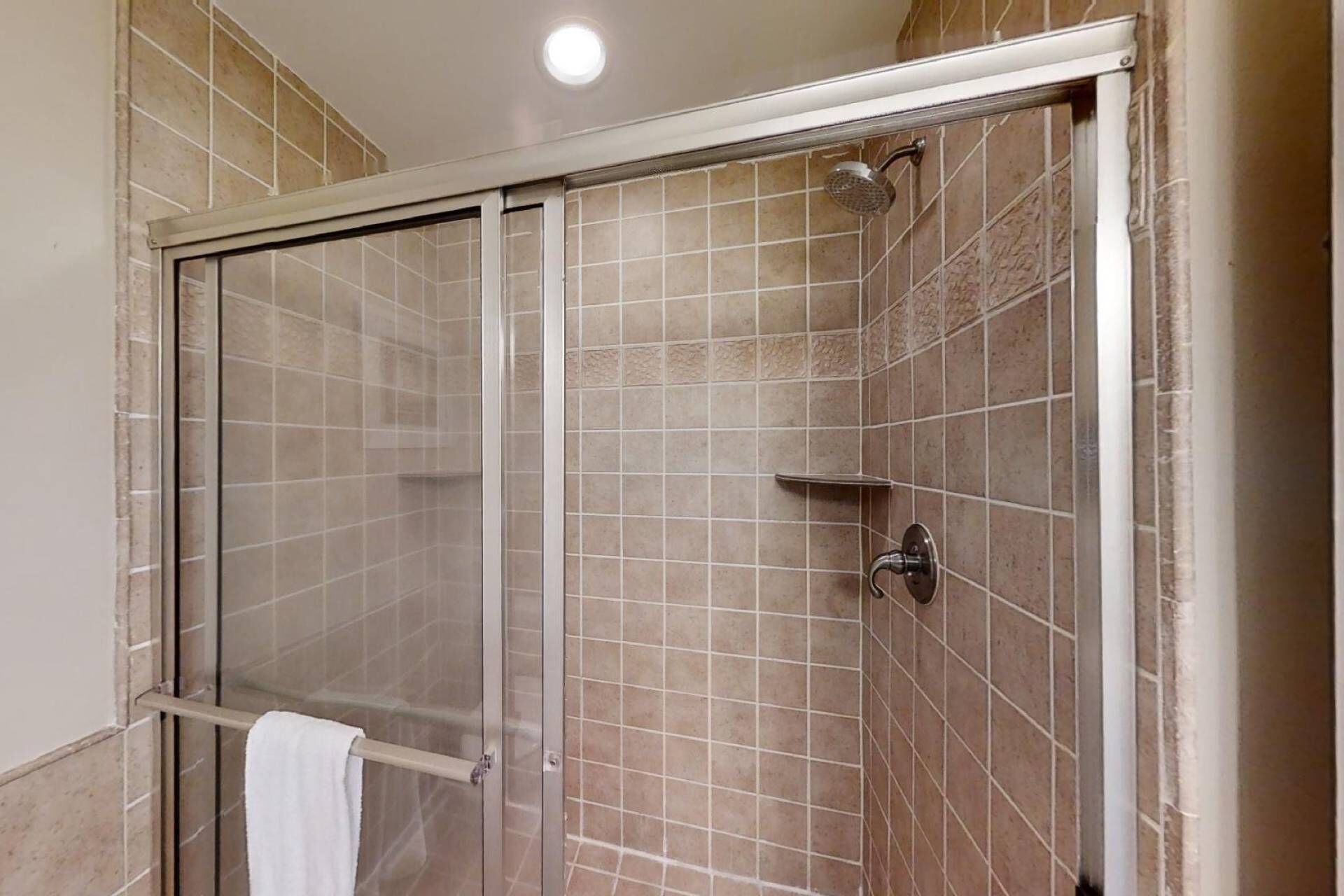 A shower with a glass door, beige tiles, and a white towel hanging on a rack.