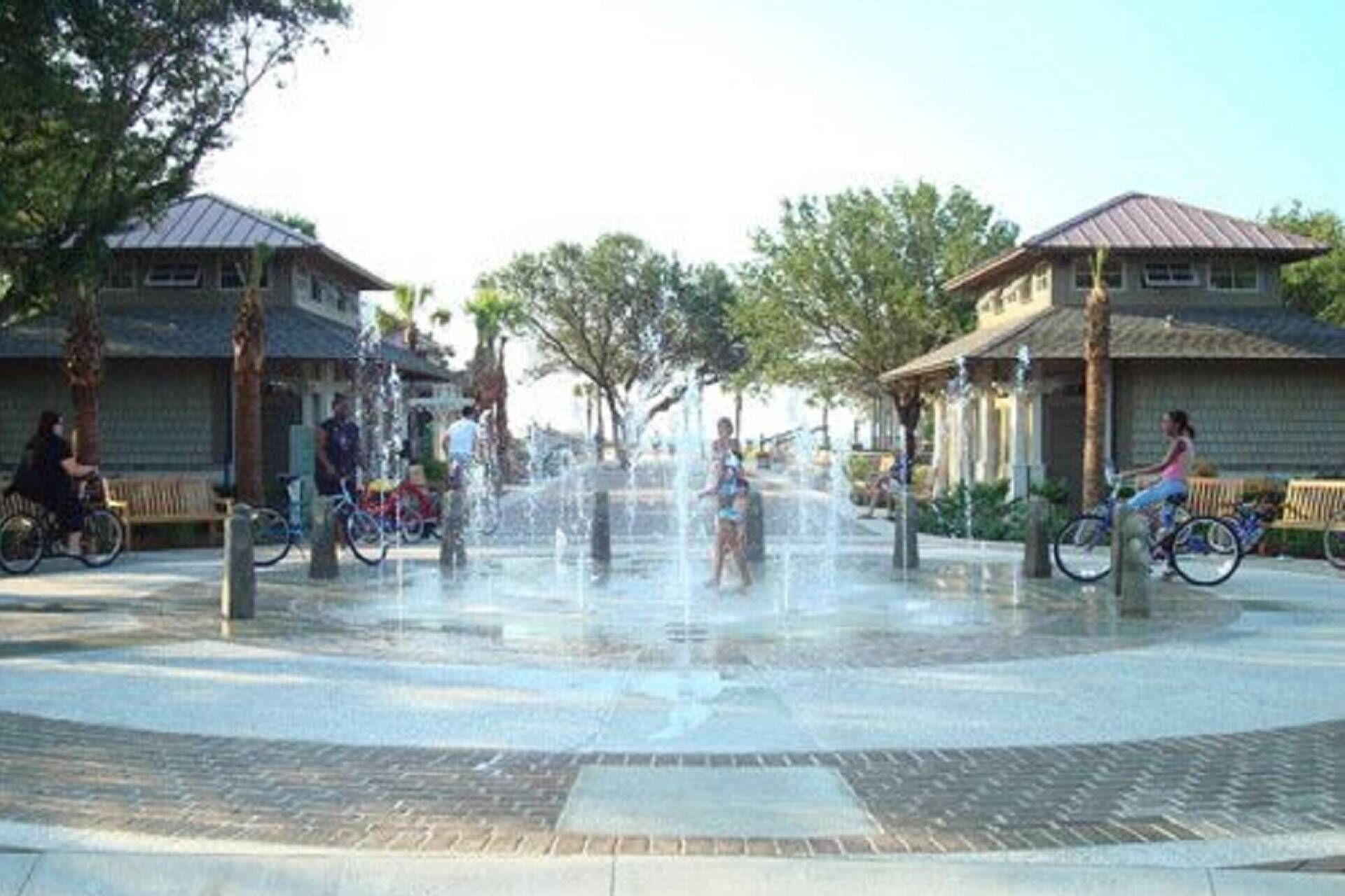 Ein öffentlicher Platz mit einem zentralen Wasserspiel, umgeben von Gebäuden mit Ziegeldächern und Bänken.