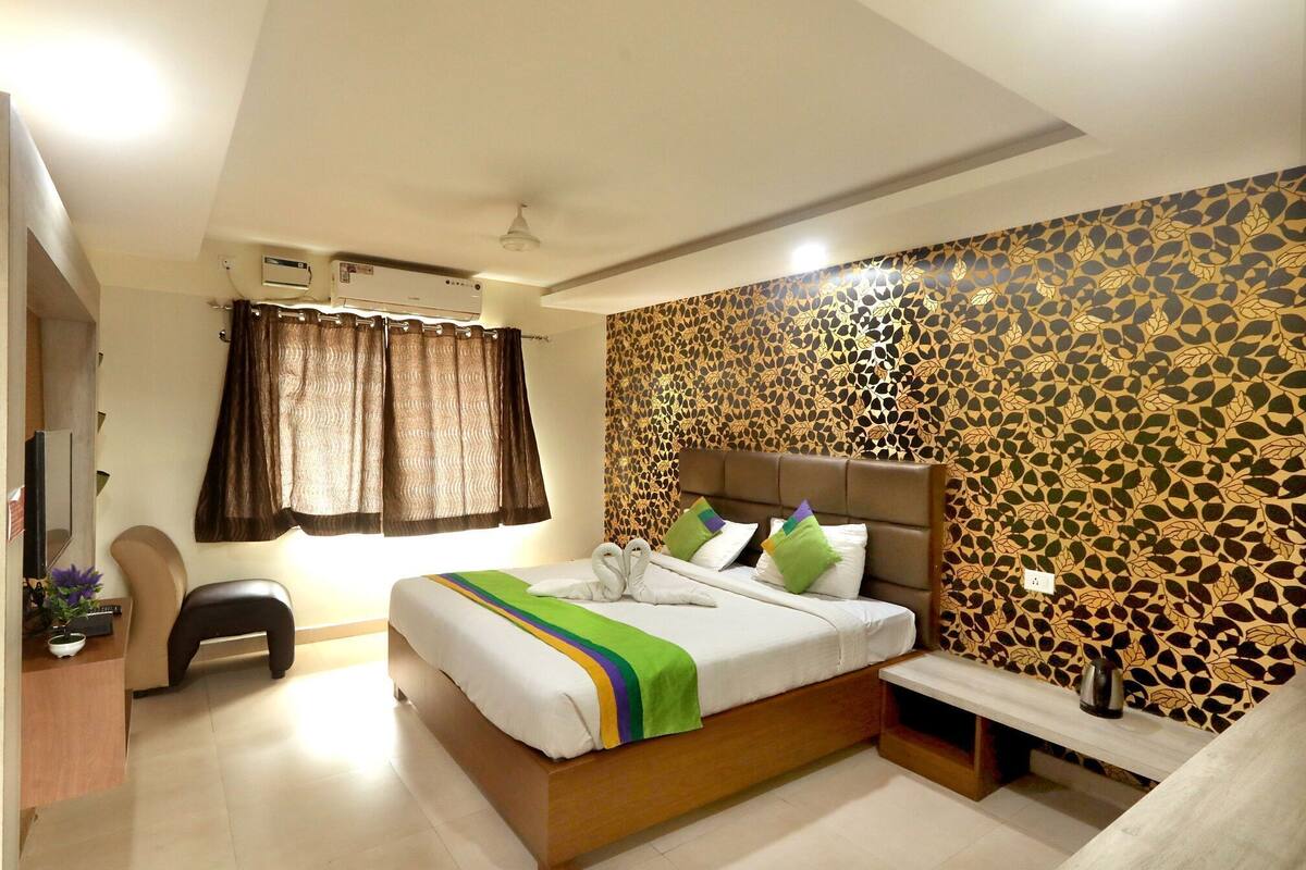 A hotel room with a bed, a desk, a chair, a window with curtains, and a patterned wall.