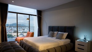Ein modernes Schlafzimmer mit einem großen Bett, einem Nachttisch und Blick auf eine Stadtlandschaft durch große Fenster.