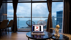 Ein modernes Zimmer mit einem Laptop auf dem Tisch, großen Fenstern mit Vorhängen und Blick aufs Meer, sowie einer kleinen Roboterfigur.