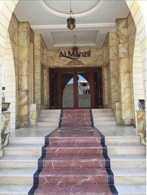 Une entrée imposante, avec des marches en marbre menant à une porte sur laquelle est inscrit le nom « Al Manzil ».
