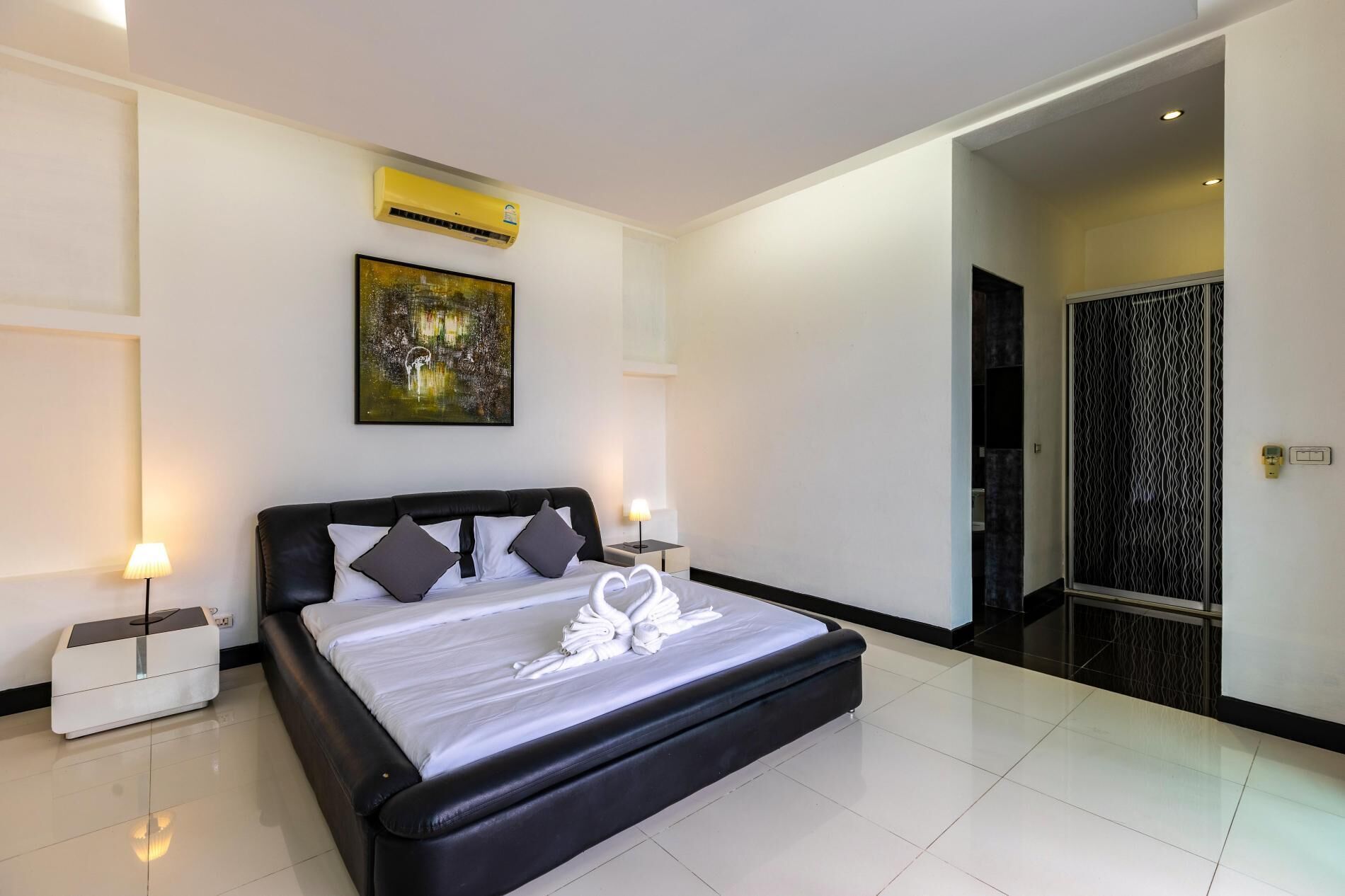 Una habitación de hotel moderna con una cama grande, un cuadro en la pared, un equipo de aire acondicionado y un baño visible a través de una puerta abierta.