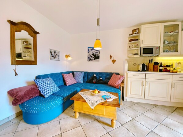 Un salon avec un canapé bleu, une table basse en bois et un coin cuisine doté de meubles blancs et d’un four à micro-ondes.