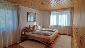 Un dormitorio con una cama de madera, dos mesitas de noche, un ventanal con cortinas ligeras y un techo con vigas de madera.