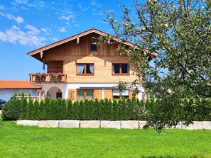 A two-story wooden house with a balcony, a stone wall in front, and a tree to the right.