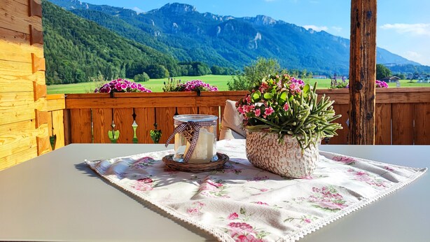 Una terraza de madera con una mesa dispuesta para dos personas, con mantel floral y una maceta con flores rosas.