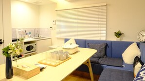 Um espaço compacto com cozinha americana, mesa de jantar com bandeja de copos, sofá com almofadas estampadas e um relógio na parede.