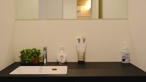 Casa de banho com bancada preta, lavatório branco, espelho, planta em vaso, dispensador de sabão, frasco de álcool em gel e garrafa de sabonete.