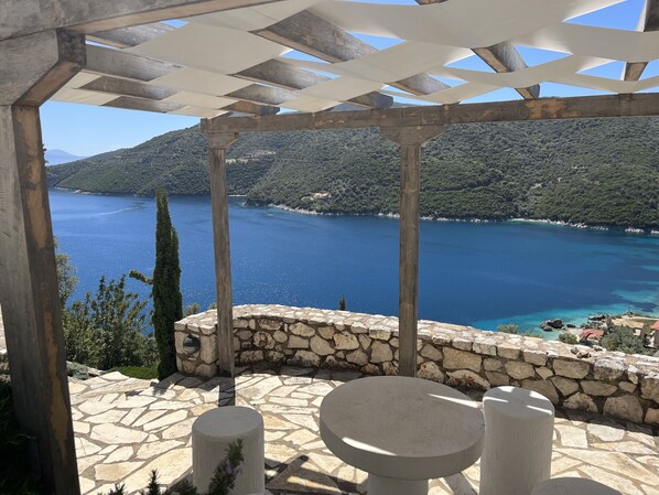 Eine terrasse mit steinbelag und einem runden Tisch sowie zwei Stühlen, mit Blick auf einen klaren, blauen See und grüne Hügel.