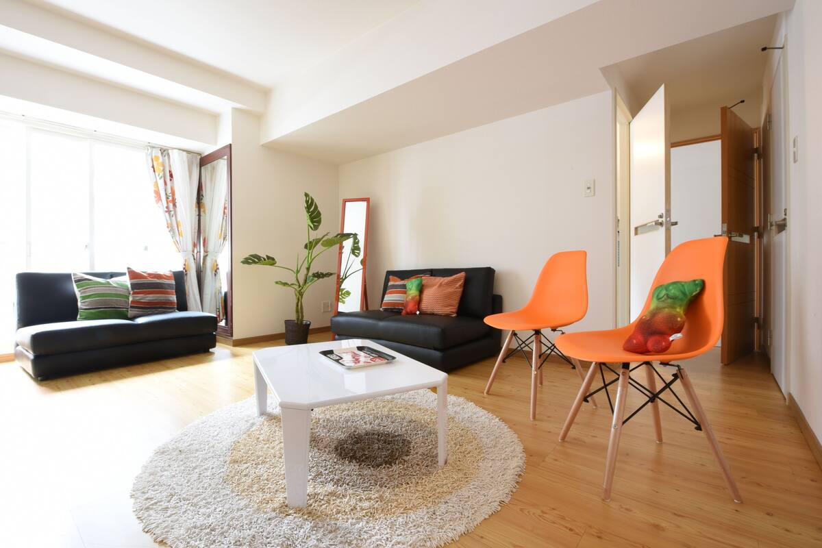 A modern living room with a sofa, a coffee table, and two chairs. There is a large window with curtains, a potted plant, and a mirror on the wall.