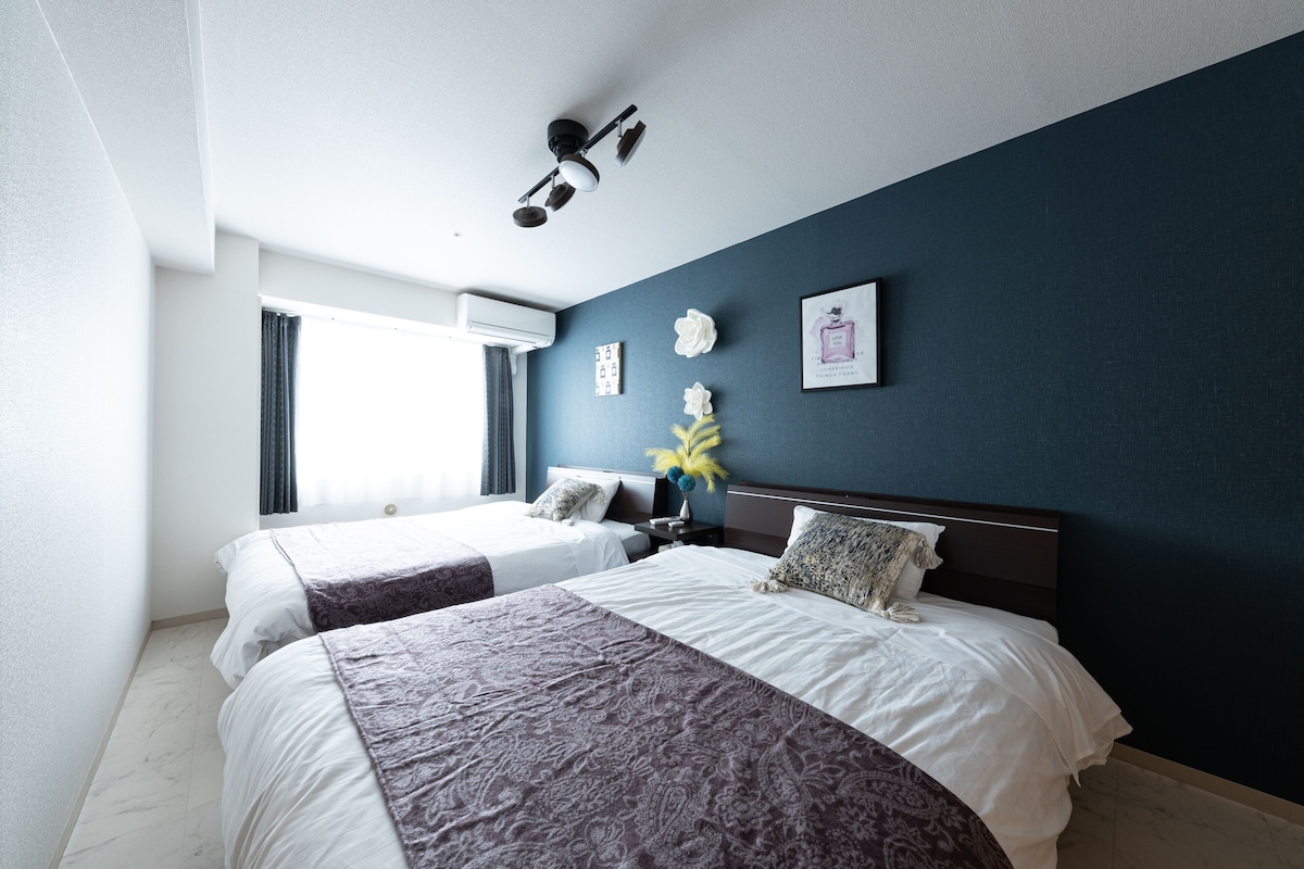 A hotel room with two beds, a ceiling fan, an air conditioning unit, a window with curtains, and a wall-mounted picture.