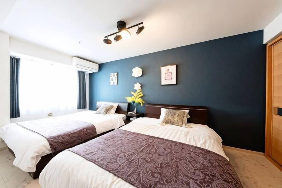 A hotel room with two beds, a dark blue accent wall, a ceiling light fixture, and a window with curtains.