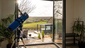 A telescope on a tripod in a room with a glass door leading to a patio with a hot tub and potted plants. - The Lee - Luxury Cottages (Keighley)