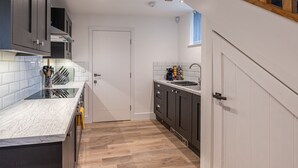 A modern kitchen with a marble countertop, wooden flooring, and dark cabinetry. - The Lee - Luxury Cottages (Keighley)