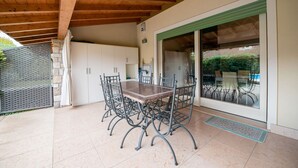 En overdækket terrasse med spisebord og stole, en stenvæg og en skydedør, der fører til et udendørs opholdsområde.